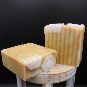 Op de afbeelding: Twee handgemaakte zeepjes, een op de ander, op een houten kruk. De zeep is lichtgeel met witte swirls.