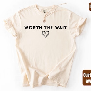 Op de afbeelding: Ivoor kleurig t-shirt met de zwarte tekst "WORTH THE WAIT" en een zwarte hart afbeelding. Het shirt heeft korte mouwen en een ronde hals. De woorden "comfort colors" en "custom text available" staan in de afbeelding.