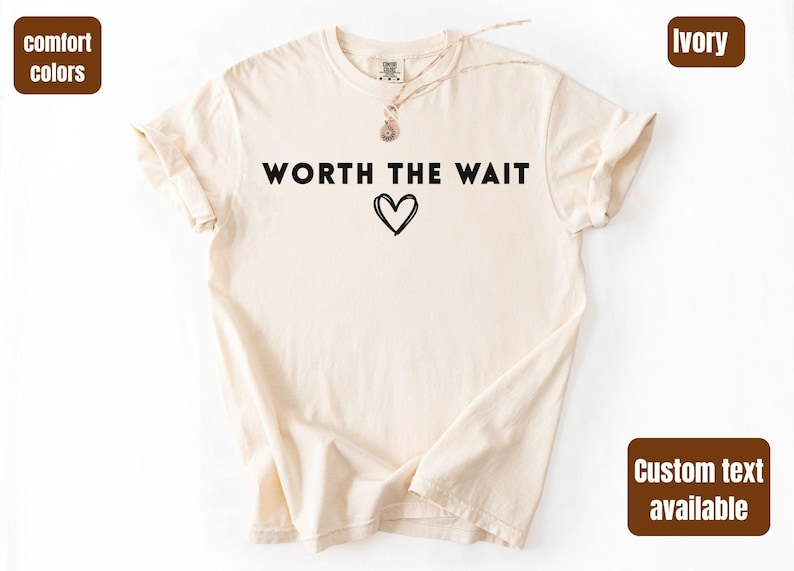 Op de afbeelding: Ivoor kleurig t-shirt met de zwarte tekst "WORTH THE WAIT" en een zwarte hart afbeelding. Het shirt heeft korte mouwen en een ronde hals. De woorden "comfort colors" en "custom text available" staan in de afbeelding.