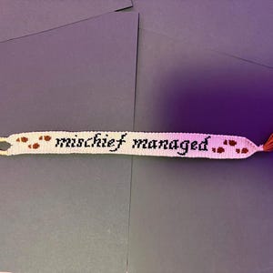 May include: A woven friendship bracelet featuring a cream-colored background with the black text "mischief managed." and small brown footprint patterns. The bracelet has a loop on one end and a short, orange-brown tassel on the other end.