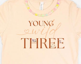 以下が含まれることがあります： 「Young Wild Three」の文字が入ったピーチ色のTシャツ、デニムショーツ、チェック柄のスリッポンシューズ、花の形をしたサングラス、ピンク色のカメラ、ビーズのネックレス。3歳児向けの楽しいコーディネート。