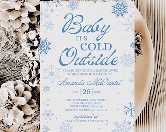 Puede incluir: Una invitación a un baby shower con el texto "Baby It's Cold Outside" en escritura azul, rodeado de copos de nieve. La invitación está sobre un fondo blanco y está decorada con piñas y vegetación.