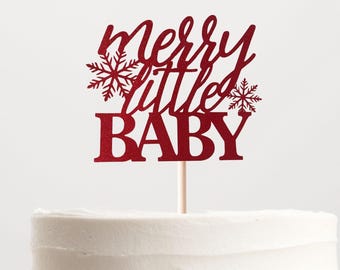 May include: A dark red cake topper featuring the text "merry little BABY" in a mix of script and block lettering, accented by two snowflake designs. The topper is attached to a thin, light-colored wooden stick and is placed on a white cake.