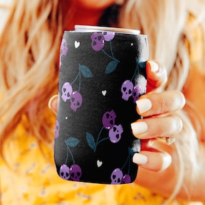 May include: Black can cooler featuring a purple skull and cherry design. The design includes purple skulls, cherries, and small white hearts on a black background. The can cooler is cylindrical and designed to keep beverages cold.