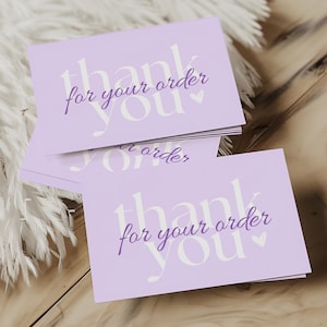 May include: A pile of pale purple cards, each bearing the words "thank you for your order" in white and purple script. They rest on a wooden surface, with white feathery decoration.