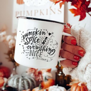 May include: White enamel mug with a black rim and handle, featuring the text "pumpkin spice & everything nice" in black script. The mug is held in front of a fall-themed display of pumpkins, gourds, and autumn leaves.