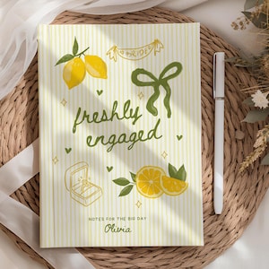 May include: A notebook with a yellow and white striped cover, featuring the text "freshly engaged" and illustrations of lemons, a ring box, and a bow. The notebook includes the text "Notes for the Big Day" and the name "Olivia".
