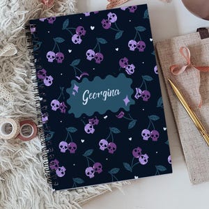 Könnte beinhalten: Ein spiralgebundenes Notizbuch mit dunkelblauem Einband und einem wiederkehrenden Muster aus violetten und hellvioletten Totenkopf-Kirschen mit grünen Stielen und kleinen weißen Herzen. Ein zentrales, wellenförmiges blaugrünes Etikett zeigt den Namen Georgina.