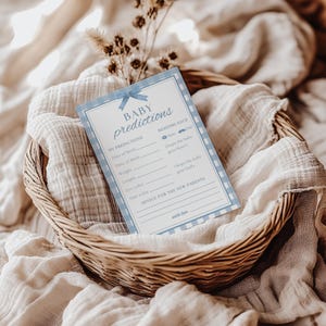 K&ouml;nnte beinhalten: Eine Babyparty-Spielkarte mit den Worten "Baby Predictions" in einem blau-wei&szlig; karierten Rand. Die Karte enth&auml;lt Abschnitte f&uuml;r Vorhersagen &uuml;ber das Geburtsdatum, das Gewicht und die Augenfarbe des Babys. Die Karte liegt in einem Weidenkorb.