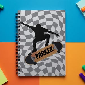 May include: Spiral-bound notebook with a black and white checkered pattern background. A black silhouette of a skateboarder is mid-air on a skateboard with the name "PARKER" on it. The notebook is on a blue and orange surface.