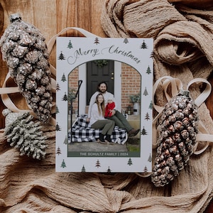 Pode incluir: Um cartão de Natal com uma foto de família, emoldurado em branco com árvores de Natal verdes e a frase «Merry Christmas». A família está sentada numa varanda. O cartão está numa bandeja de madeira com decorações de pinhas.