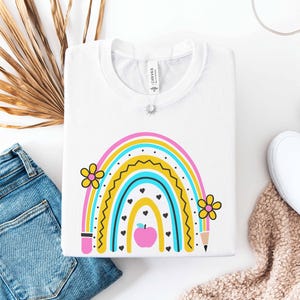 May include: White crew neck t-shirt featuring a colorful rainbow design with flowers, hearts, and an apple. The rainbow includes pink, yellow, blue, and black colors. A pencil and coffee cup are also part of the design. The shirt is folded on a white surface.