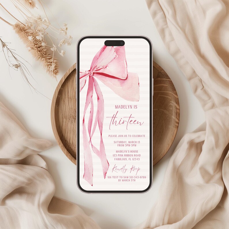 May include: A smartphone displays a pink bow invitation for a thirteenth birthday. The invitation reads "Madelyn is thirteen" with event details. The phone rests on a wooden plate, with beige fabric in the background.