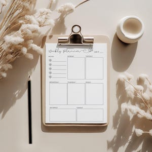 May include: A wooden clipboard with a weekly planner sheet, featuring sections for each day and notes. The planner is held by a silver clip. A black pencil and a small white ceramic bowl are also present.