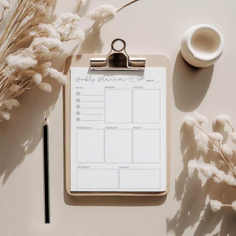 May include: A wooden clipboard with a weekly planner sheet, featuring sections for each day and notes. The planner is held by a silver clip. A black pencil and a small white ceramic bowl are also present.
