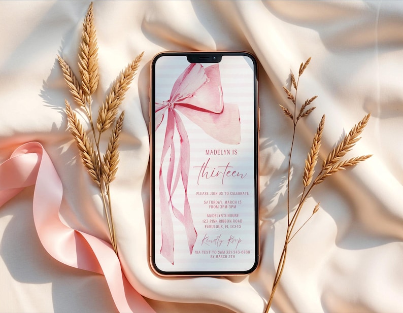 May include: A smartphone displays a pink watercolor bow and the text "Madelyn is thirteen" on a white background. The phone is surrounded by wheat stalks and a pink ribbon on a satin surface.