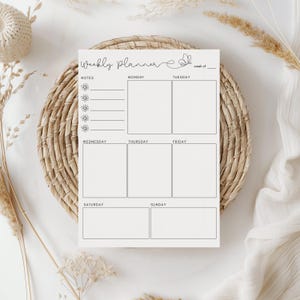 May include: A cream-colored weekly planner with the words "Weekly Planner" in cursive script and a butterfly graphic. The planner has sections for notes and daily scheduling, resting on a woven placemat.