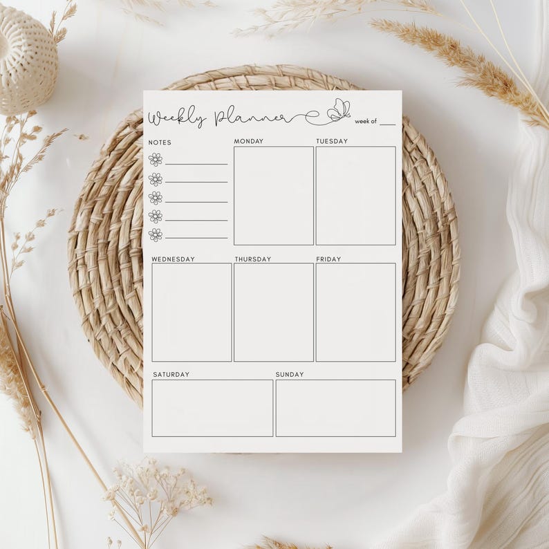 May include: A cream-colored weekly planner with the words "Weekly Planner" in cursive script and a butterfly graphic. The planner has sections for notes and daily scheduling, resting on a woven placemat.