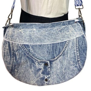 May include: A blue denim shoulder bag with a white trim and a patterned strap. The bag has a buttoned front design and a zipper closure. The strap is blue and white with a geometric pattern.