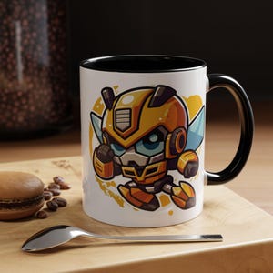 May include: A white ceramic mug with a black rim and handle. The mug features a cartoon illustration of a yellow and black robot with wings. The robot has a blue face and is in a flying pose.