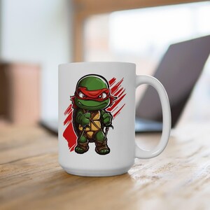 May include: White ceramic mug with a cartoon image of a Teenage Mutant Ninja Turtle wearing a red bandana and holding two katanas. The turtle is green with a yellow shell and brown straps. The background is white with red brushstrokes.