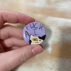 May include: A round, purple button with the text "FUCK A.I." in black. The button features a cartoon cat with black and tan markings. The cat has a grumpy expression.