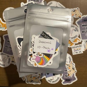 Puede incluir: Dos bolsas de plástico transparente que contienen paquetes de pegatinas. Un paquete tiene una etiqueta que dice "Oopsies! 10-Pack" con una flor morada. Se ven pegatinas con varios diseños, incluido un caracol arcoíris.