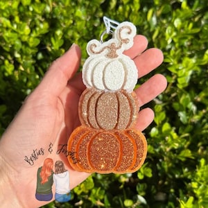 May include: A hanging ornament featuring a stack of three pumpkins in descending order of size. The top pumpkin is white, the middle is copper, and the bottom is orange. Each pumpkin is covered in a textured glitter finish with a metal loop.