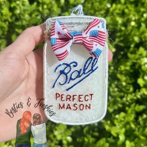 May include: A white mason jar-shaped ornament with the text "Ball PERFECT MASON" in blue and red glitter. It has a bow with a red, white, and blue American flag pattern. The ornament is held against a green background.