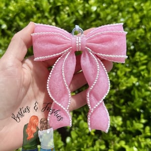 May include: A pink textured bow ornament featuring a white beaded trim along the edges and a silver metal loop at the top for hanging. The text Besties & Freshies is printed on the lower left along with an illustration of two people.