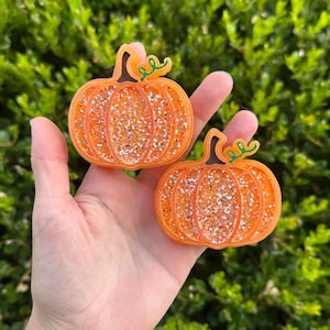 Puede incluir: Dos decoraciones en forma de calabaza naranja con purpurina y zarcillos verdes. Las calabazas se sostienen en una mano sobre un fondo de follaje verde. Decoraciones para otoño o Halloween.