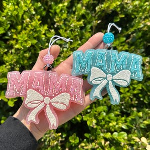 May include: Two hanging ornaments shaped like the word MAMA with a white bow graphic. One ornament is pink with glitter, and the other is light blue with glitter. Each features a small beaded loop at the top for hanging purposes.