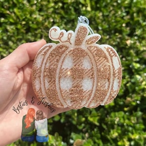 May include: A pumpkin-shaped air freshener featuring a white base with a rose gold glitter buffalo plaid pattern. The item includes a small white loop at the top for hanging and displays the text Besties & Freshies with a cartoon illustration.