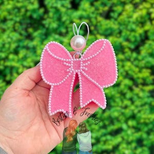 May include: A bright pink textured bow ornament featuring a white pearl bead border and a single large white pearl bead at the top. A white elastic loop is attached to the top for hanging. The text Besties & Freshies is visible at the bottom.