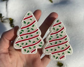 Puede incluir: Dos adornos con forma de árbol de Navidad. Cada adorno tiene un contorno blanco, un centro transparente y está decorado con rayas rojas y puntos verdes. Los adornos se sostienen en una mano sobre un fondo nevado.