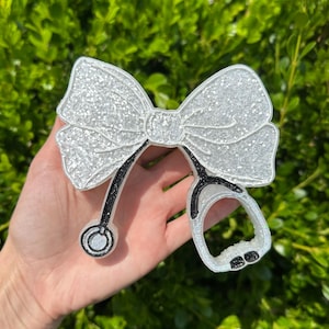 May include: A decorative bow ornament featuring a white glitter finish with a black glitter stethoscope design integrated into the bottom. The bow has a textured surface and a central knot detail, with the stethoscope tubing extending downwards.