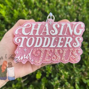 May include: A pink, glittery, resin ornament with the words "CHASING TODDLERS and JESUS" in white script. The ornament is held in a hand against a blurred green background. The words "Besties & Freshies" are also visible.