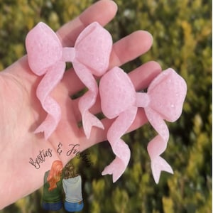 May include: Two light pink resin bows with a glittery texture. The bows have long, flowing tails and are held in a hand. The bows are a craft item and are labeled with the text "Besties & Freshies".
