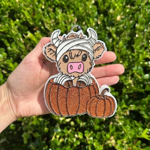 May include: A decorative ornament featuring a brown Highland cow wrapped in white bandages like a mummy, sitting inside a large, glittery orange pumpkin next to a smaller matching glittery orange pumpkin, with a metal loop at the top.