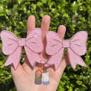 May include: Two pink glittery bow-shaped hair clips. The bows are a light pink color and have a sparkly glitter finish. The image also includes a small illustration of two people sitting together.