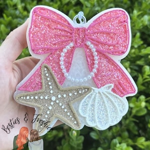 K&ouml;nnte beinhalten: Ein dekorativer Lufterfrischer in Form einer gro&szlig;en, glitzernden rosa Schleife mit einer wei&szlig;en perlenartigen Schlaufe, einem beigen, strukturierten Seestern und einer wei&szlig;en, schillernden Muschel. Oben befindet sich eine kleine Metall&ouml;se.