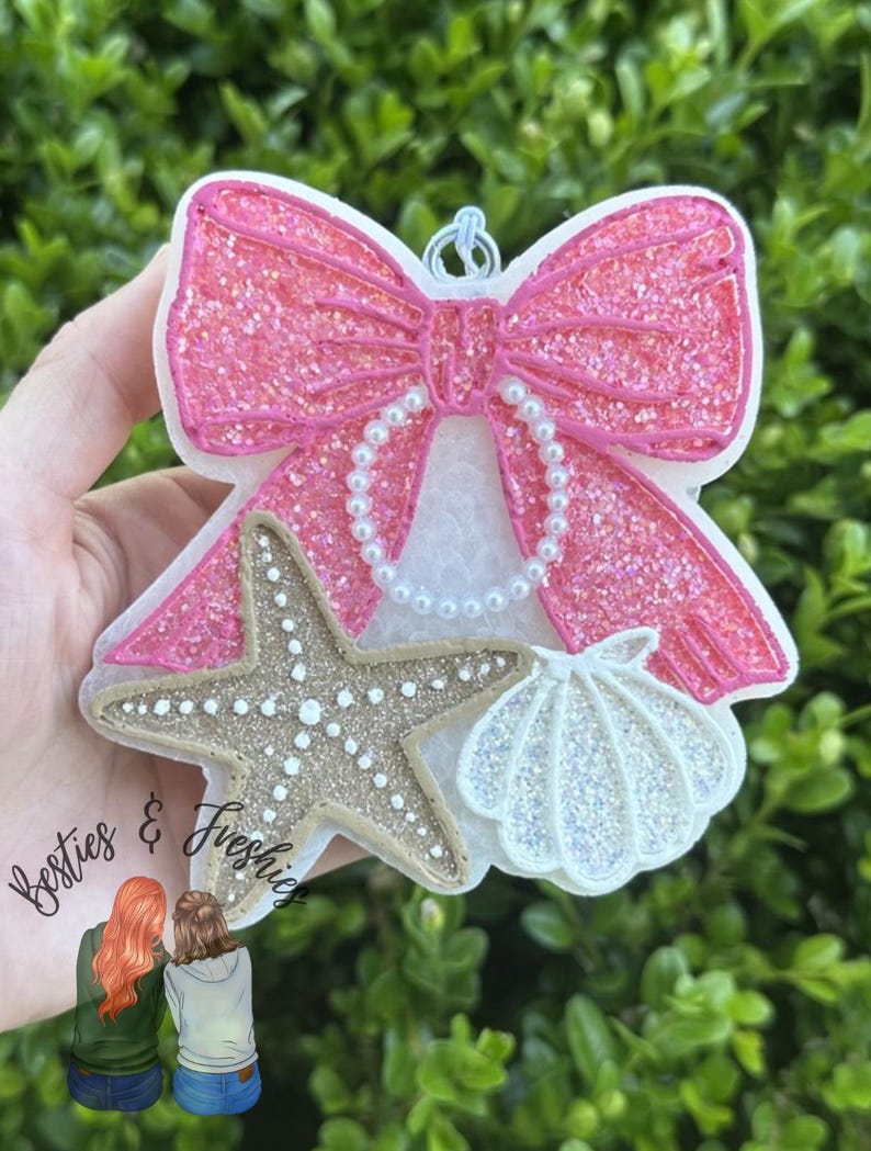 K&ouml;nnte beinhalten: Ein dekorativer Lufterfrischer in Form einer gro&szlig;en, glitzernden rosa Schleife mit einer wei&szlig;en perlenartigen Schlaufe, einem beigen, strukturierten Seestern und einer wei&szlig;en, schillernden Muschel. Oben befindet sich eine kleine Metall&ouml;se.