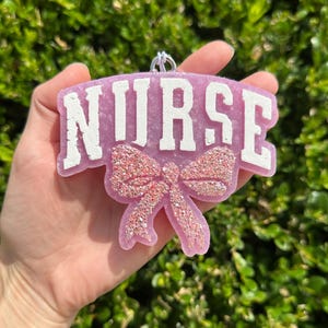May include: A lavender-colored resin ornament shaped like a curved plaque with the word NURSE embossed in white block letters. Below the text is a prominent, textured pink glitter bow. A small silver metal loop is attached to the top center.