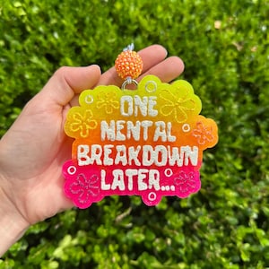 Może przedstawiać: Kolorowy brelok z białym napisem "ONE MENTAL BREAKDOWN LATER...". Brelok ma gradient żółtego, pomarańczowego i różowego z kwiatowymi akcentami. Mały, pomarańczowy koralikowy urok wisi na górze.
