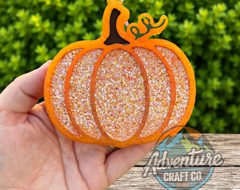 Puede incluir: Un adorno con forma de calabaza que presenta un borde naranja brillante y un tallo marrón oscuro. Las secciones interiores están rellenas con una capa densa de purpurina iridiscente multicolor que brilla. Un pequeño lazo blanco está unido al tallo.