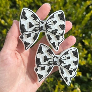 May include: Two white and black cow print bows. The bows have a glittery black outline and black spots. The bows are held in a hand against a blurred green background.
