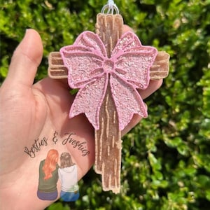 May include: A decorative cross with a pink glitter bow. The cross is a light brown color with a textured surface. The bow is a light pink color with glitter. The cross is held in a hand, with the text "Besties & Freshies" and an illustration of two women.