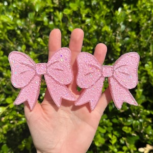 Puede incluir: Dos pinzas para el pelo con lazo rosa brillante. Los lazos están hechos de un material translúcido y están decorados con purpurina iridiscente. Los lazos se sostienen en una mano sobre un fondo de follaje verde.