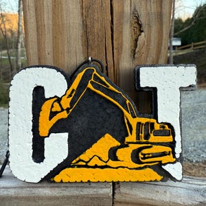 May include: A three-dimensional wall hanging featuring the letters C and T in white with a textured surface. Between the letters is a black background with a yellow excavator and a pile of dirt, creating the CAT logo design with a hanging loop.