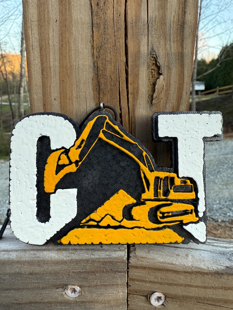 May include: A three-dimensional wall hanging featuring the letters C and T in white with a textured surface. Between the letters is a black background with a yellow excavator and a pile of dirt, creating the CAT logo design with a hanging loop.